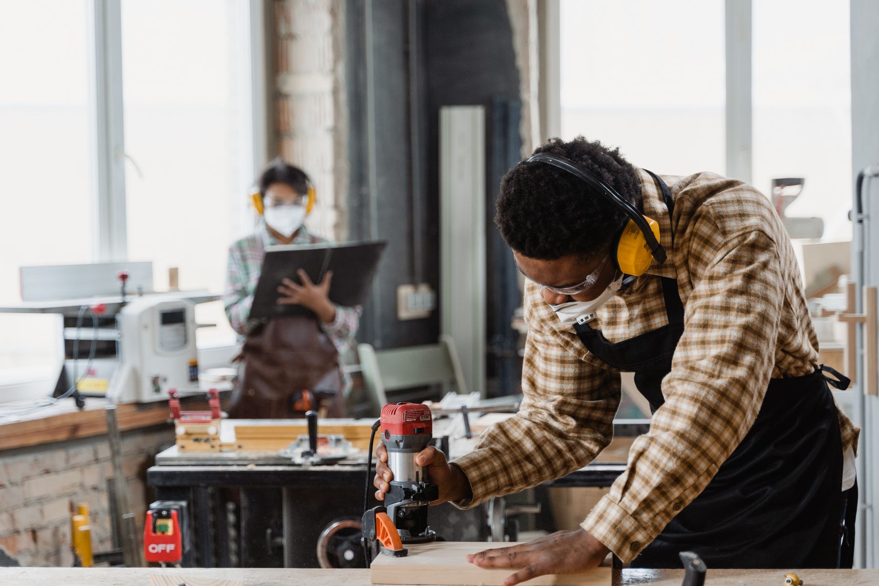 A carpenter using a woodworking tool with a colleague in the workshop